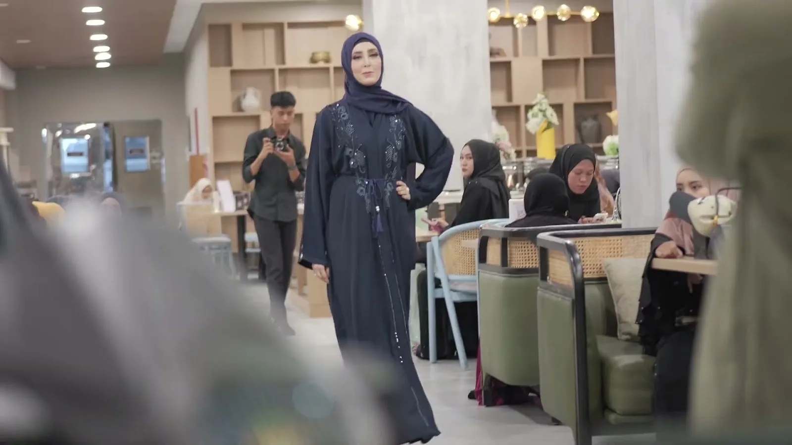 Model mengenakan busana Heelwa berwarna biru gelap di area fashion show dengan pengunjung di sekelilingnya